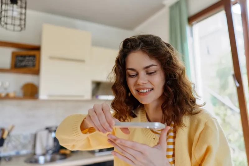 Smiling woman in a yellow sweater prepares cooking ingredients in a bright kitchen, measuring and pouring. Cozy home lifestyle scene with natural light and a warm, relaxed mood and calm