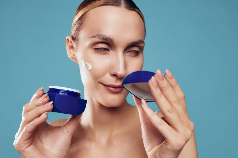 Woman skincare face cream beauty routine, holding a blue jar and applying moisturizer with spatula, eyes closed relaxed smile, spa mood, soft smooth skin, blue background