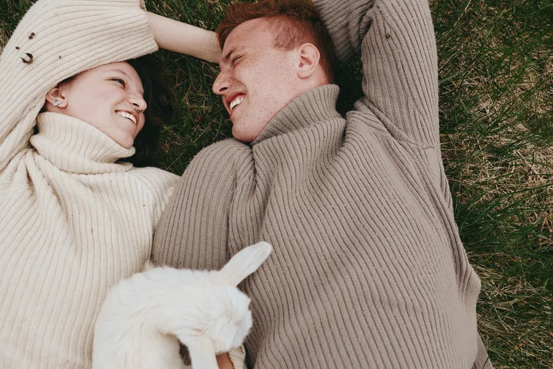 Joyful couple outdoors, lying on grass with a playful goat kid nearby, cozy sweaters, sunlit scene, relaxed mood, candid affection, natural colors, serene countryside vibe