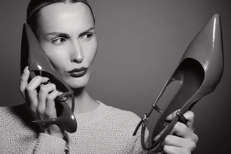 White woman with pale skin and dark lipstick holds pointed shoes near her face looking thoughtfully to the side. Concept of fashion accessories and style choices in monochrome.