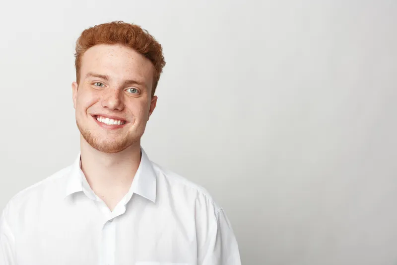 Smiling young man with red hair and beard wearing white shirt stands against plain light background. Portrait of happy confident male with casual style and natural expression.