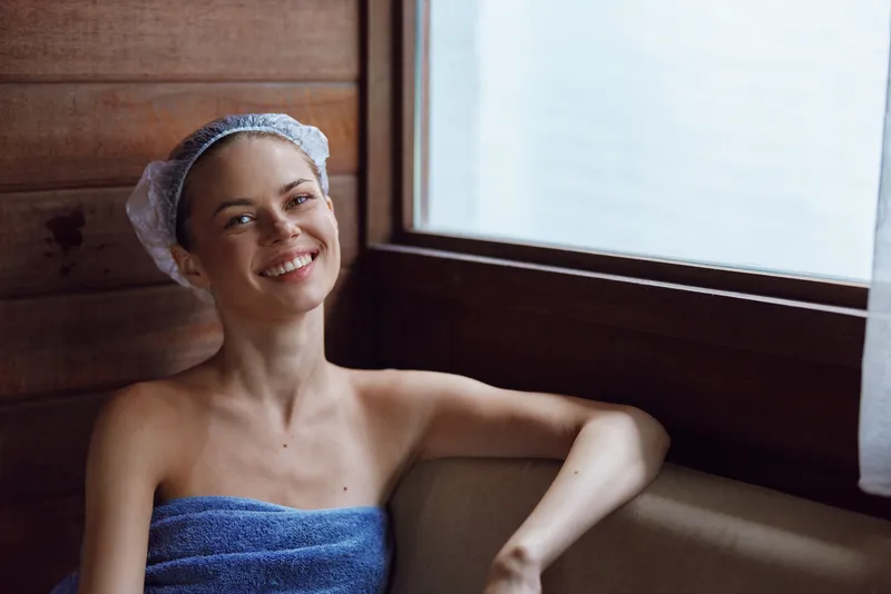 smiling woman wrapped in blue towel with hair cap relaxing indoors by wooden wall near large window, natural light, cozy spa atmosphere
