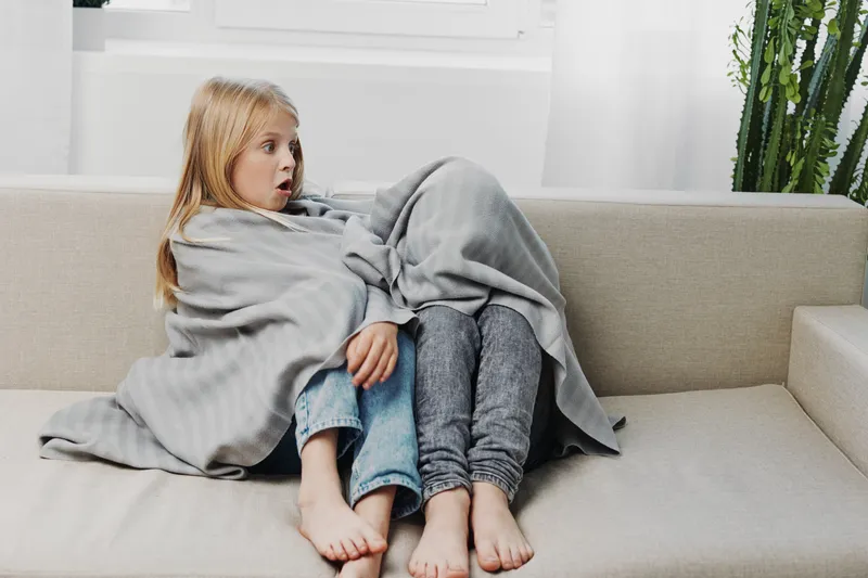 Two surprised children wrapped in a blanket sit closely on a beige sofa, watching something off camera in a bright living room with natural light.