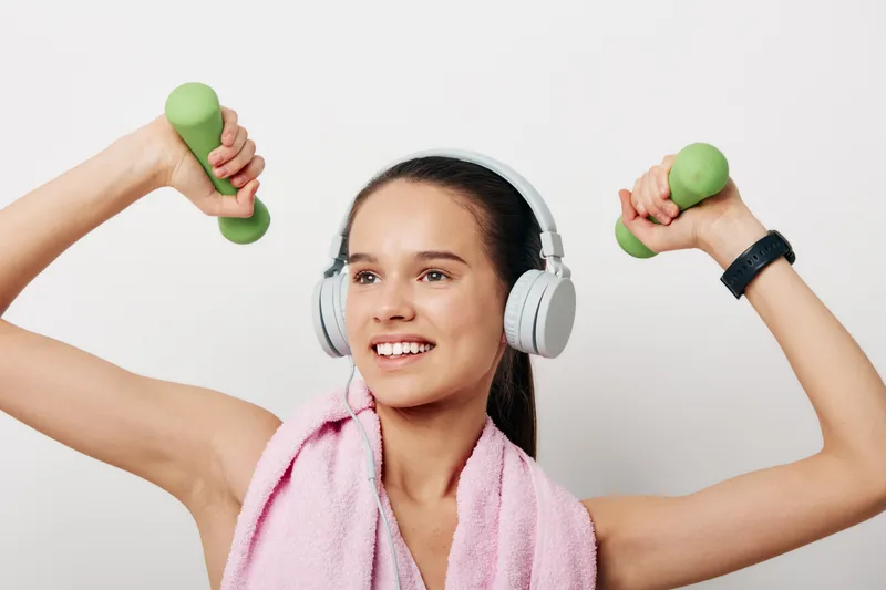 Active woman in a bright studio lifting small green dumbbells, wearing headphones and a towel around her neck, mid workout showcasing fitness, sport and training energy.
