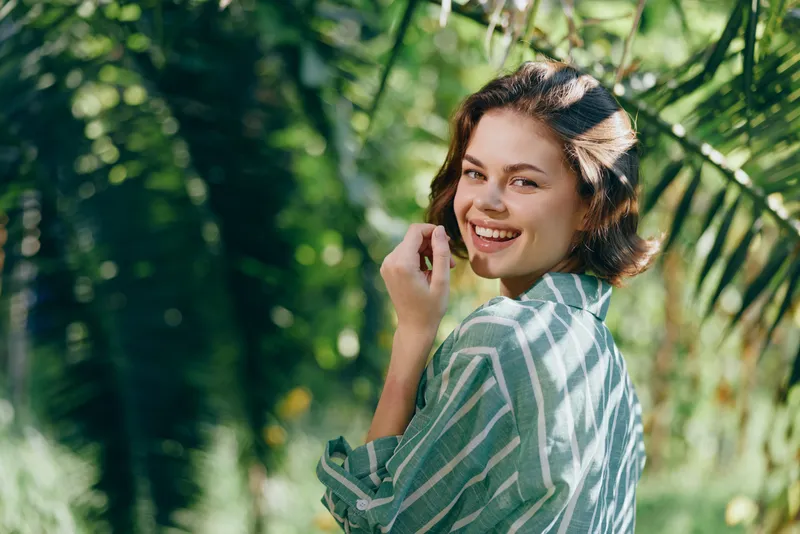 Smiling woman outdoors in a striped shirt turns toward the camera in a sunlit garden, creating a fresh, natural casual portrait.