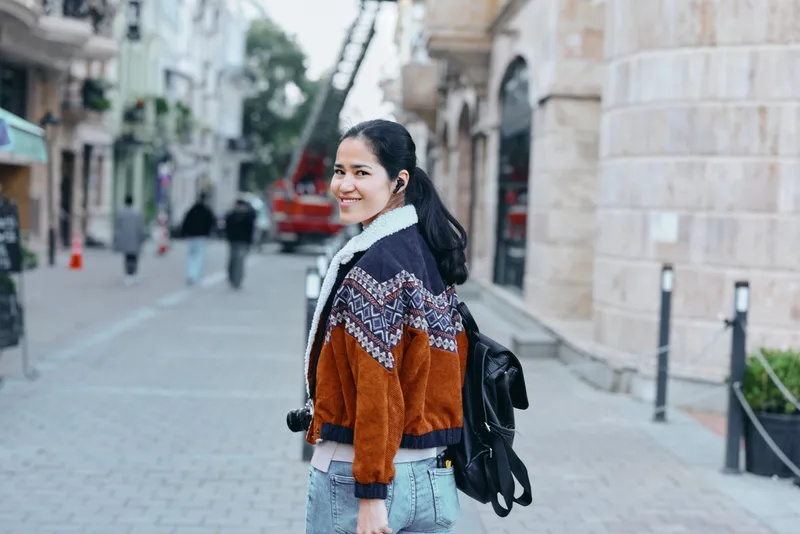 Woman with backpack walking along an urban street, wearing a colorful knit jacket, denim jeans and scarf, exploring a sunlit avenue with stone buildings and pedestrian traffic and distant storefronts.