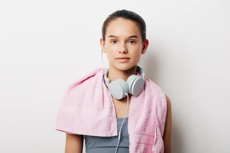 Woman in athletic gear with a towel over her shoulders and headphones, ready for a workout in a studio setting. Clean, focused portrait highlighting fitness lifestyle and sport discipline.