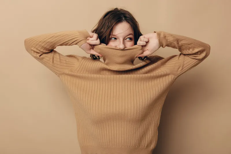 Young woman in brown knitted sweater pulls collar over her mouth, looking sideways with playful expression. Studio portrait on beige background with casual fashion and cozy mood.