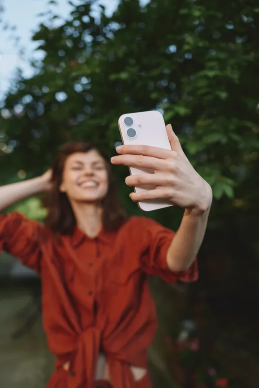 Young woman takes a selfie outdoors, wearing a red casual shirt, smiling and relaxed, capturing a lifestyle portrait against a green park backdrop