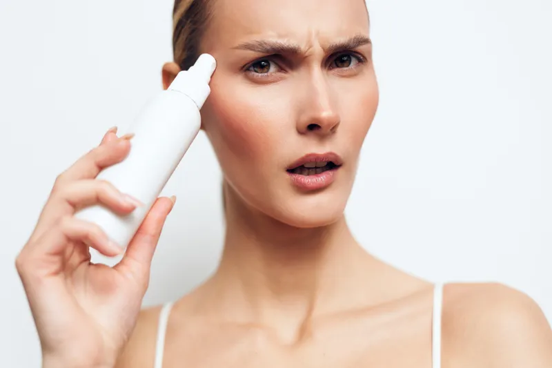A concerned woman holds a white spray bottle near her face, showing a puzzled expression. The image represents skincare routine, beauty, and personal care concepts.