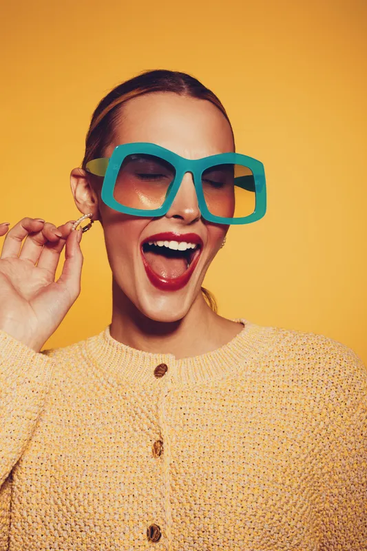 Happy woman with large turquoise sunglasses and yellow sweater laughs joyfully on yellow background. Concept of fashion, summer style, and positive emotions in studio lighting.
