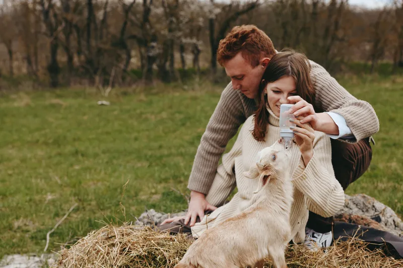 Casual couple outdoors in a park share a tender moment with a light colored dog, wearing warm knitwear and sipping a drink, creating a cozy lifestyle scene of affection and companionship