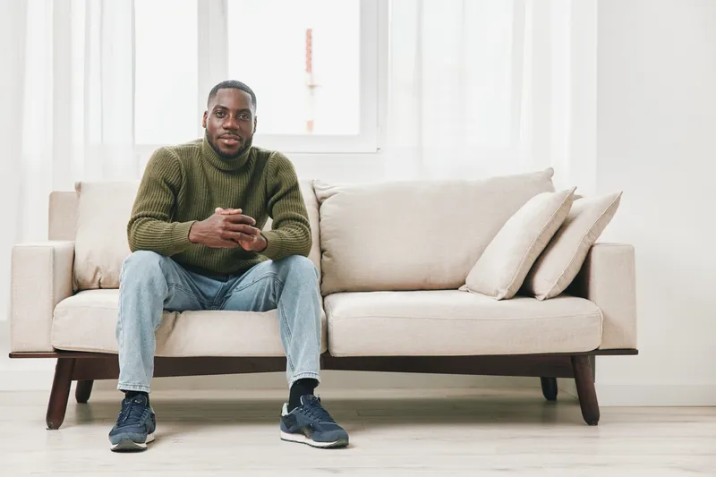 Young Black man relaxing on a beige sofa, wearing a green sweater and jeans, using a smartphone in a bright, modern living room