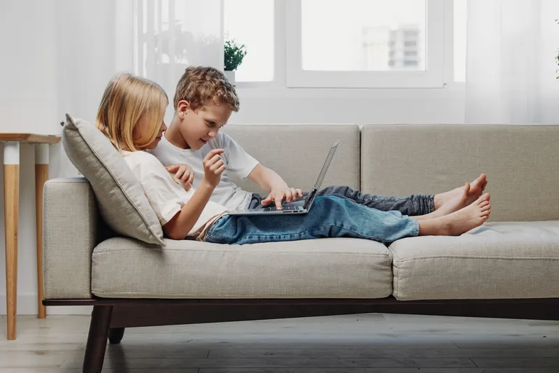 Two children lying on a modern sofa using a laptop together in a bright living room. Casual home environment with natural light, showing technology use, learning, and sibling bonding.