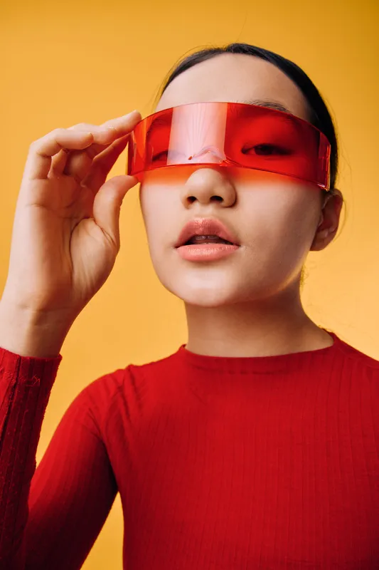 Woman with red transparent visor on face and red sweater posing on yellow background. Beauty portrait with modern futuristic fashion style and minimalistic colorful studio shot.