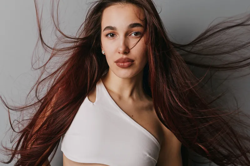 Woman with long flowing hair and natural makeup posing against gray background. Female model wearing white top with confident expression and loose hair movement in studio light.