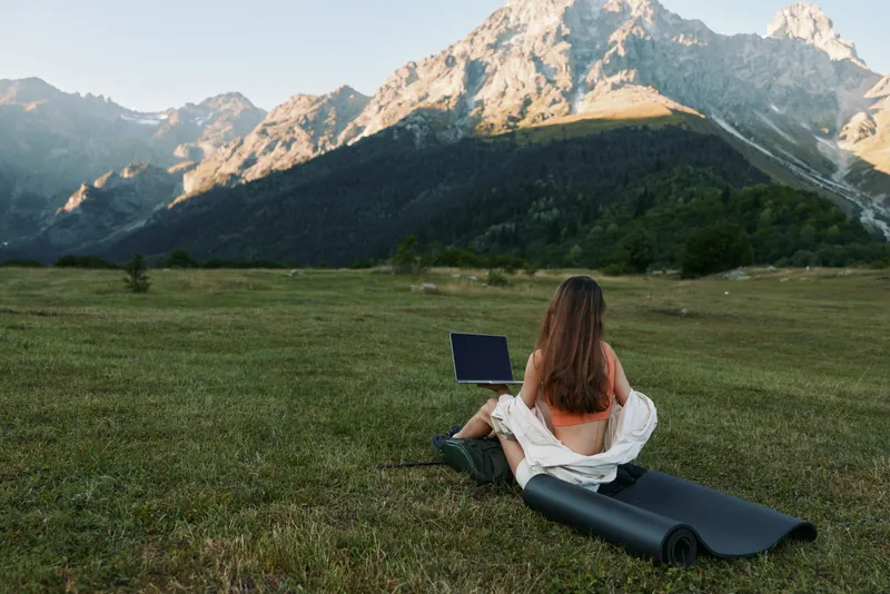 A woman sits on a grassy field with a laptop, framed by rugged mountains. She wears casual clothes, appears focused and relaxed, creating a peaceful outdoor work atmosphere.