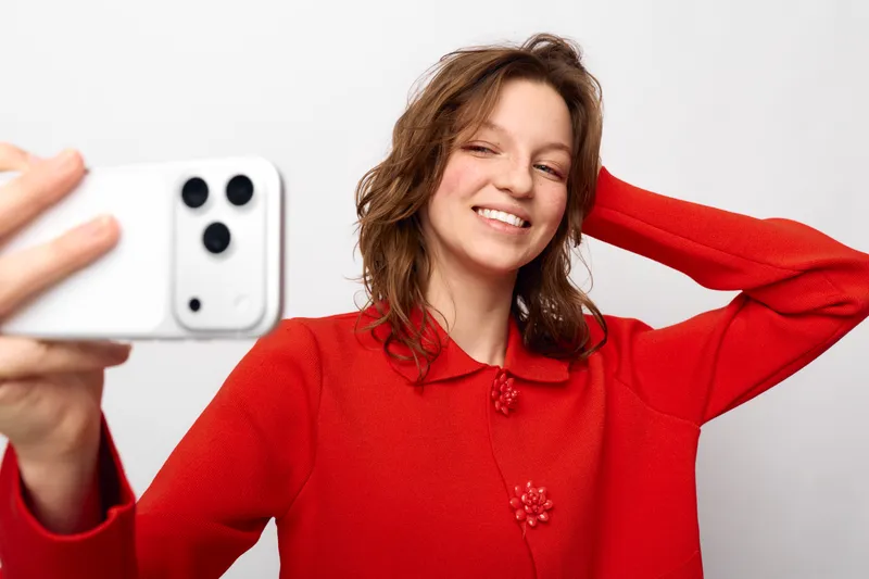 Smiling woman in red sweater takes selfie with smartphone on white background. Casual lifestyle, happiness, and modern technology concept with natural light and relaxed pose.