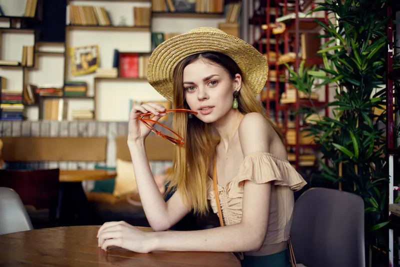 Симпатичная женщина в шляпе сидит за столом с книгами на заднем плане, крупный план