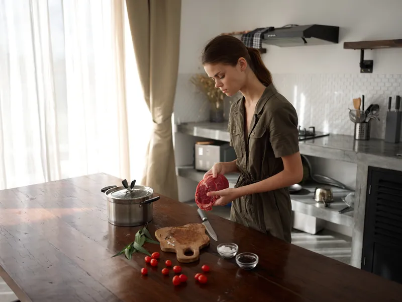 Женщина на кухне готовит ужин из мяса, овощей и специй на деревянном столе на фоне стильной кухни
