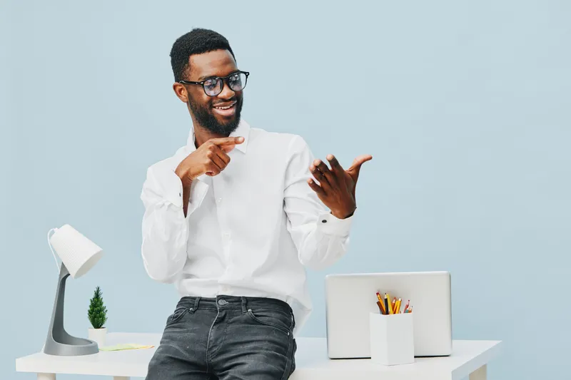 Smiling man with glasses in white shirt standing at modern workspace, pointing at smartphone, cheerful expression, casual office environment, light blue background, professional lifestyle concept, day