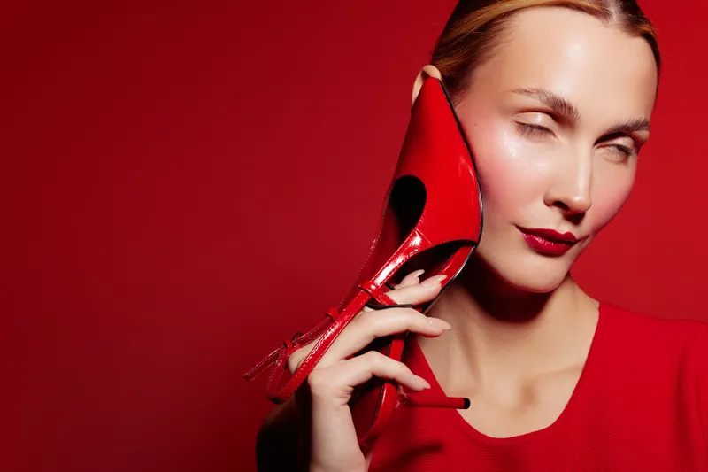 Woman in red dress holds red high heel shoe near face on red background. Fashion concept with bold makeup and stylish accessories in studio lighting.
