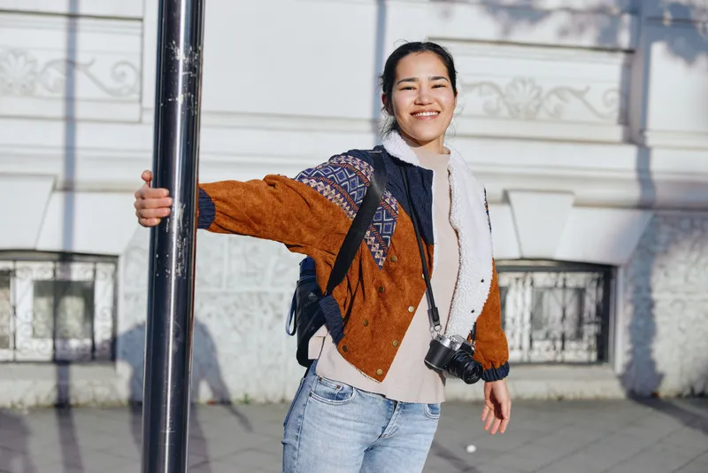 Smiling woman with backpack and camera on a street corner, outdoor fashion in urban casual style. A candid moment showcasing streetstyle, denim, scarf, and daylight for lifestyle photography in city
