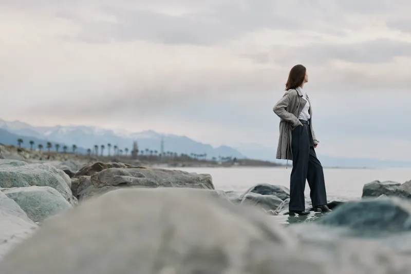 A contemplative woman stands on rocky beach looking at ocean horizon, wearing casual clothes. Overcast sky and distant mountains create calm and reflective mood in nature setting.