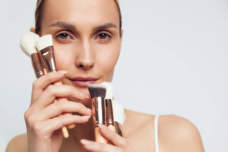 A young woman with natural skin holds several makeup brushes close to her face, showcasing beauty tools in a bright studio setting. Concept for skincare and cosmetics.