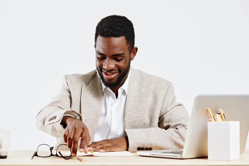 Professional young African American man working at desk in modern office, using laptop computer and glasses, smiling, dressed in beige blazer and white shirt, isolated on plain white background, busin