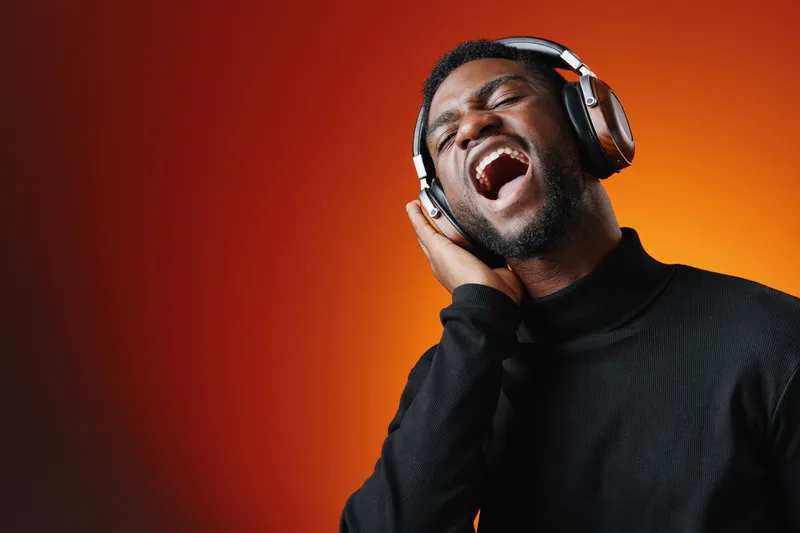 A young Black man enjoys music passionately while wearing headphones, expressing vibrant emotions against a warm gradient background