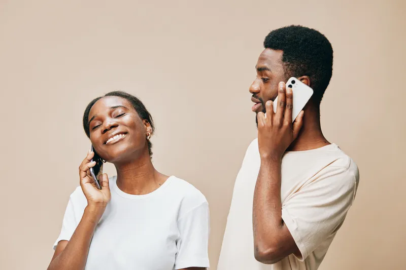 Two young Black adults joyfully talking on their smartphones, showcasing modern communication and connection in a warm, neutral toned background