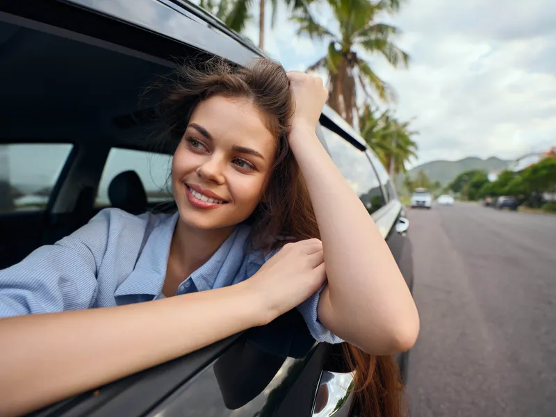 Улыбающаяся женщина выглядывает из окна автомобиля на фоне пальм, запечатлевая момент радости и свободы под солнечным небом