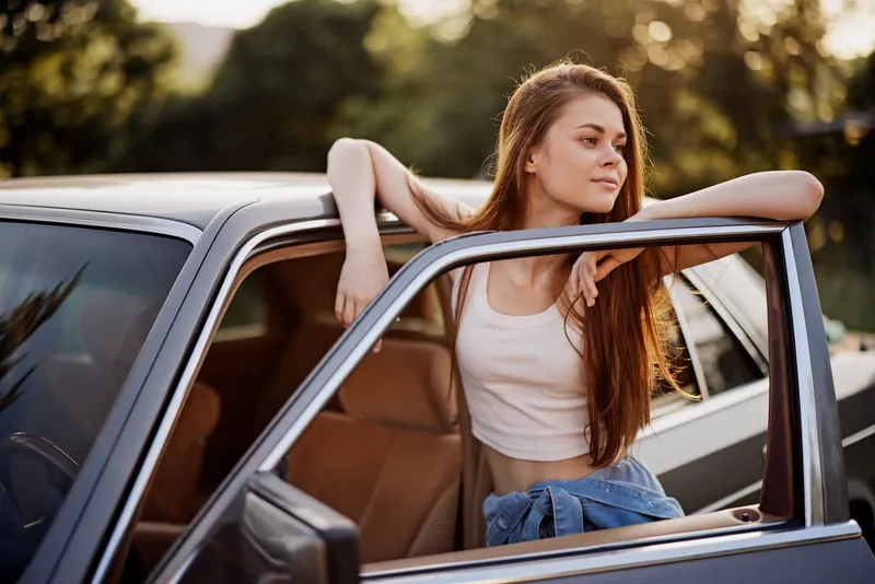 Young woman leaning on a car door in a summer setting, wearing a casual outfit, expressing relaxed joy, with lush green background and warm sunlight.