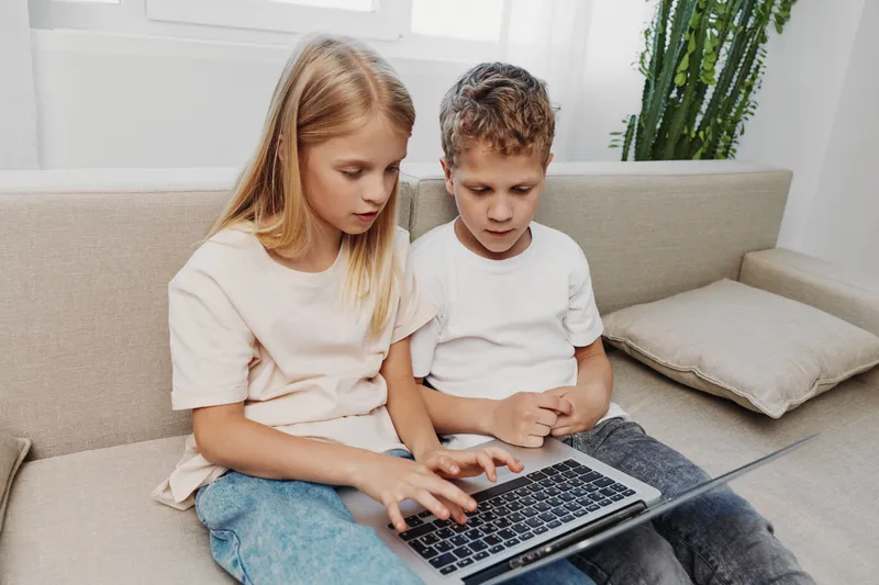 Two focused children sitting on sofa using laptop together in bright room. Concept of teamwork, learning, technology and childhood development with casual home atmosphere.