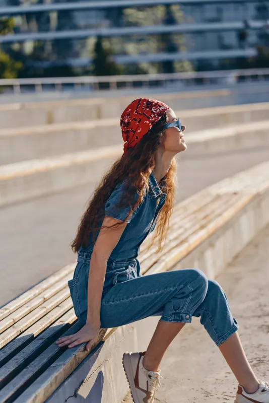 Lifestyle woman in modern boho-western denim street style sitting outdoors wearing a red bandana and sunglasses with film color effect, enjoying sunny urban atmosphere.