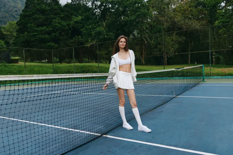 Young female tennis player in a sporty outfit, smiling confidently on an outdoor tennis court, surrounded by greenery and natural light
