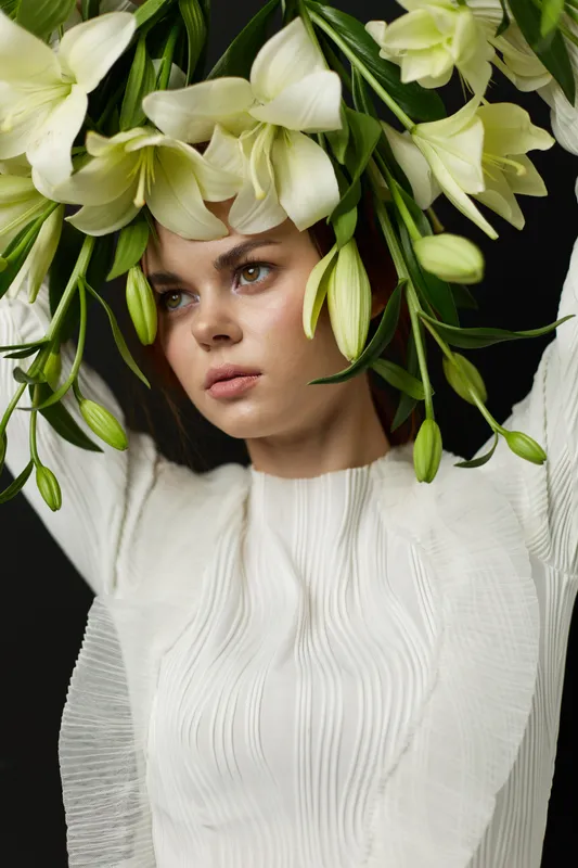 A young woman with a serene expression adorned with a floral headpiece of white lilies, wearing a textured white dress against a dark background