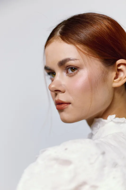Elegant portrait of a young woman with red hair in a white vintage dress, conveying confidence and serenity on a soft, neutral background
