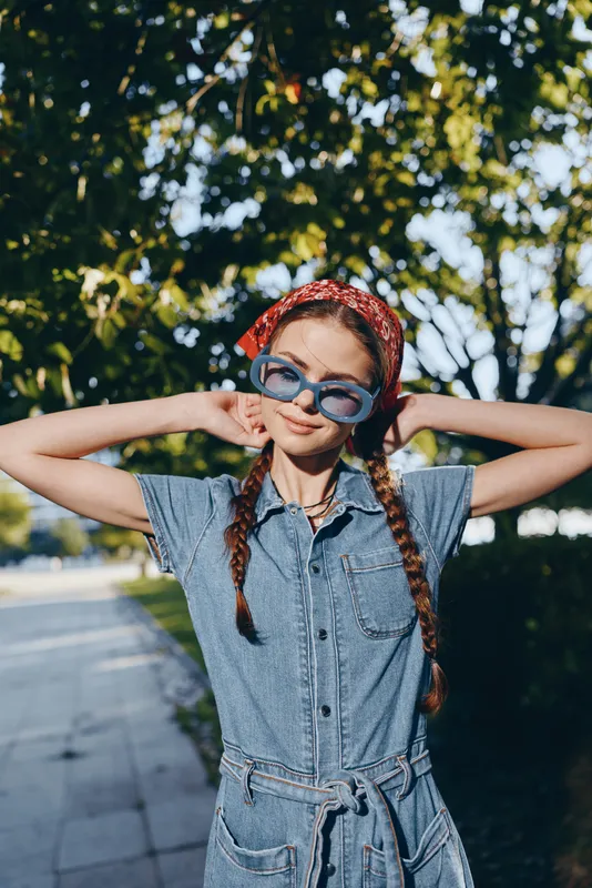 young woman wearing sunglasses and denim dress with red bandana smiles outdoors in bright sunny day with green trees in background