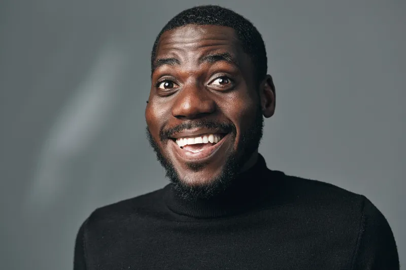 African man with dark skin smiling and wearing black turtleneck sweater in creative conceptual visual style. Portrait of happy male with short hair and beard on gray background.