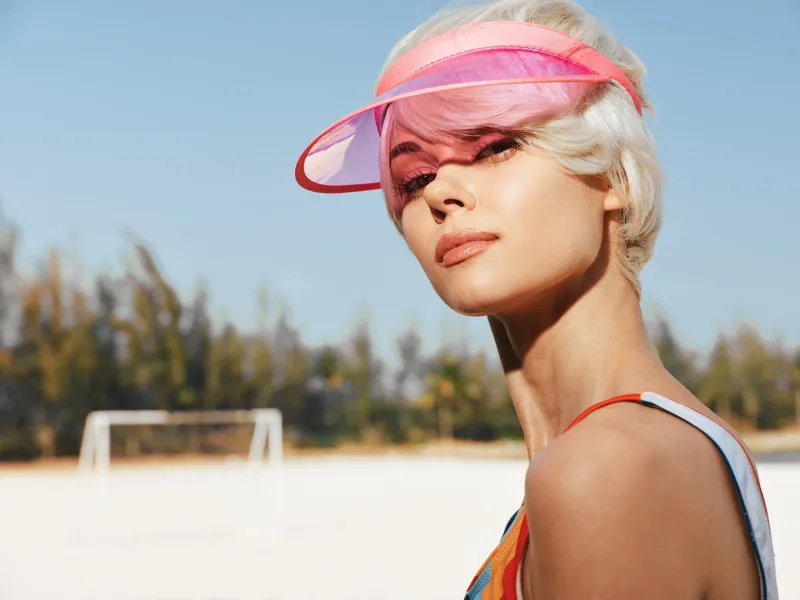 Portrait of a stylish woman wearing a pink visor on a sunny beach. Bright light highlights sun kissed skin, bold makeup, and a relaxed summer mood with outdoor vibe.