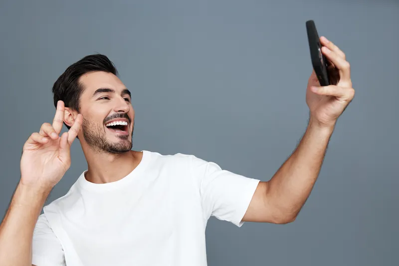 Happy smiling young man taking selfie with smartphone, wearing white t-shirt, standing against solid grey background, enjoying moments of joy and connection.