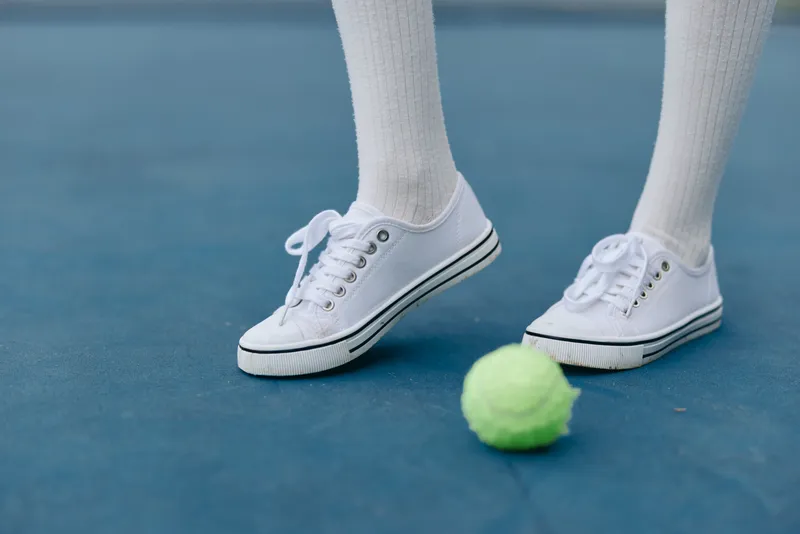 A close up of sporty white sneakers on a blue tennis court, showcasing athletic footwear with vibrant green tennis ball, perfect for sports enthusiasts and summer activities
