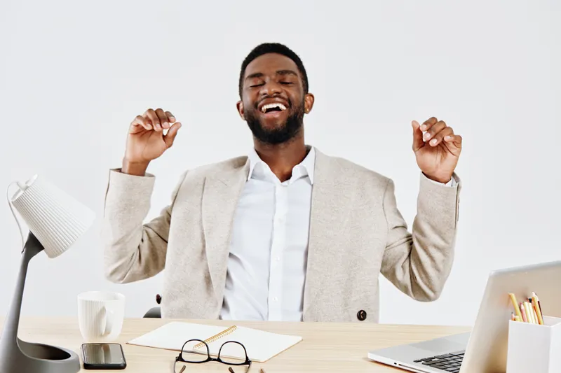 Smiling happy young African American man in beige blazer and white shirt enjoying success at work siting at modern office desk with laptop, coffee mug, eyeglasses and stationery. People lifestyle conc