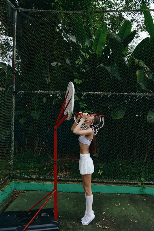 Young female athlete practicing basketball in sporty white outfit against a lush green background, showcasing determination and focus on the court