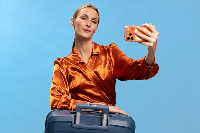 Young woman in orange satin blouse takes selfie with smartphone while holding blue suitcase on blue background. Concept for travel, vacation, and social media lifestyle.