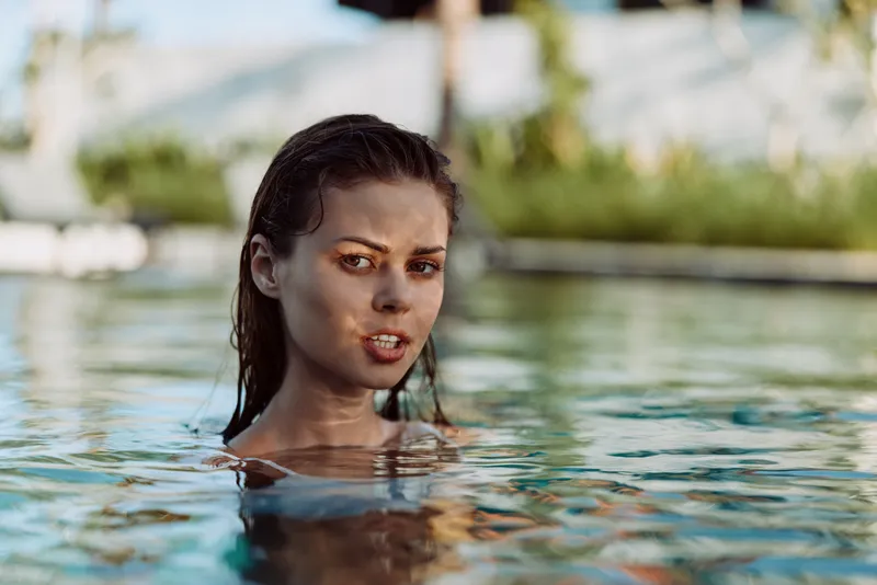 Woman in a pool with shoulders above water, facing the camera with a calm look, outdoors. Sunlit scene conveys leisure, wellness, and relaxed summer lifestyle.