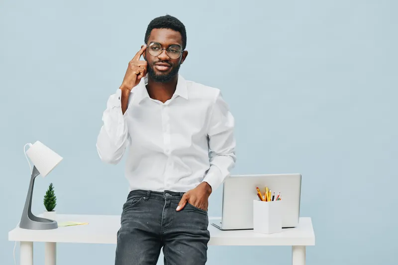 Confident young African American man in white shirt standing near desk with laptop and stationery, posing thoughtfully against plain light blue background for business or technology concept.