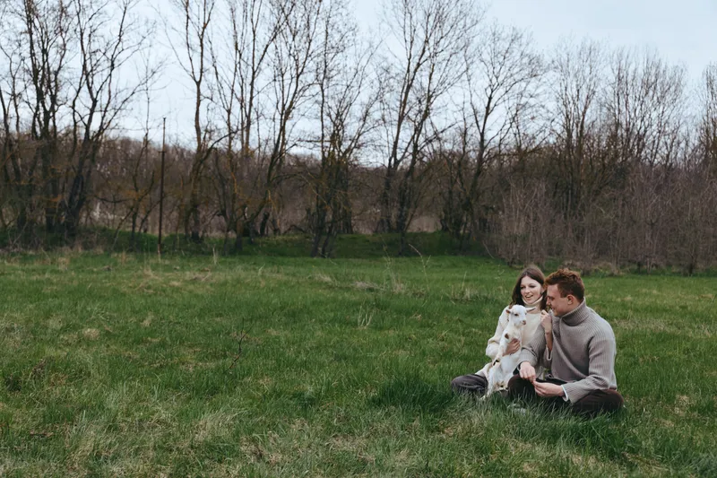Casual couple sits on a grassy field with their dog, enjoying a relaxed countryside moment, wearing sweaters and smiling softly, warm natural light, peaceful outdoors, simple companionship and joy